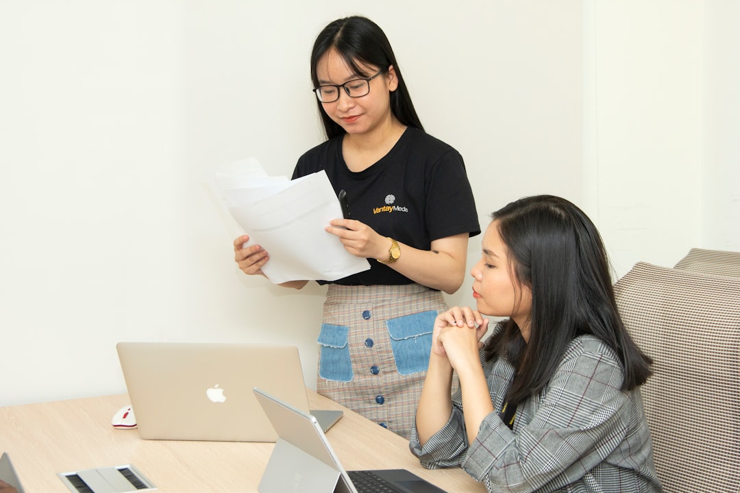 Two women working at a table with laptops; one is standing and reading papers, the other is seated and listening.