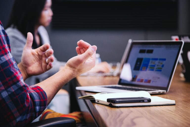 Person gesturing during a meeting with an open laptop, notebook, and phone on the conference table.