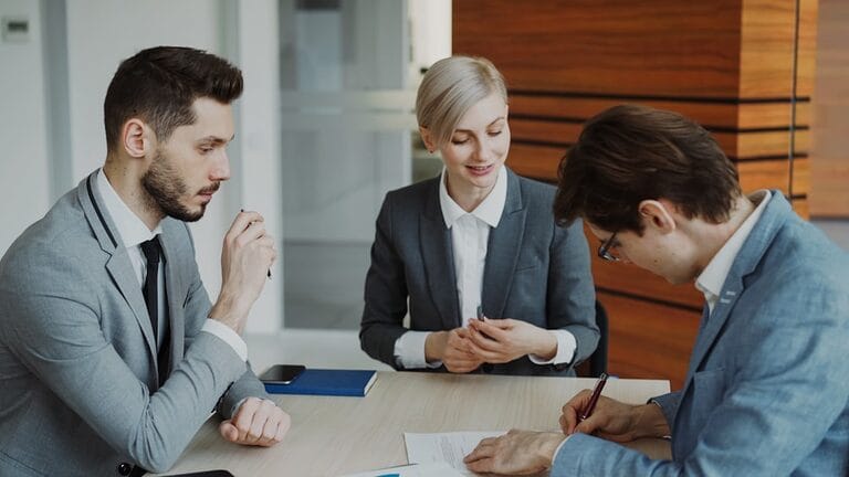 Three people in business attire sit at a table; one is signing a document while others look on.