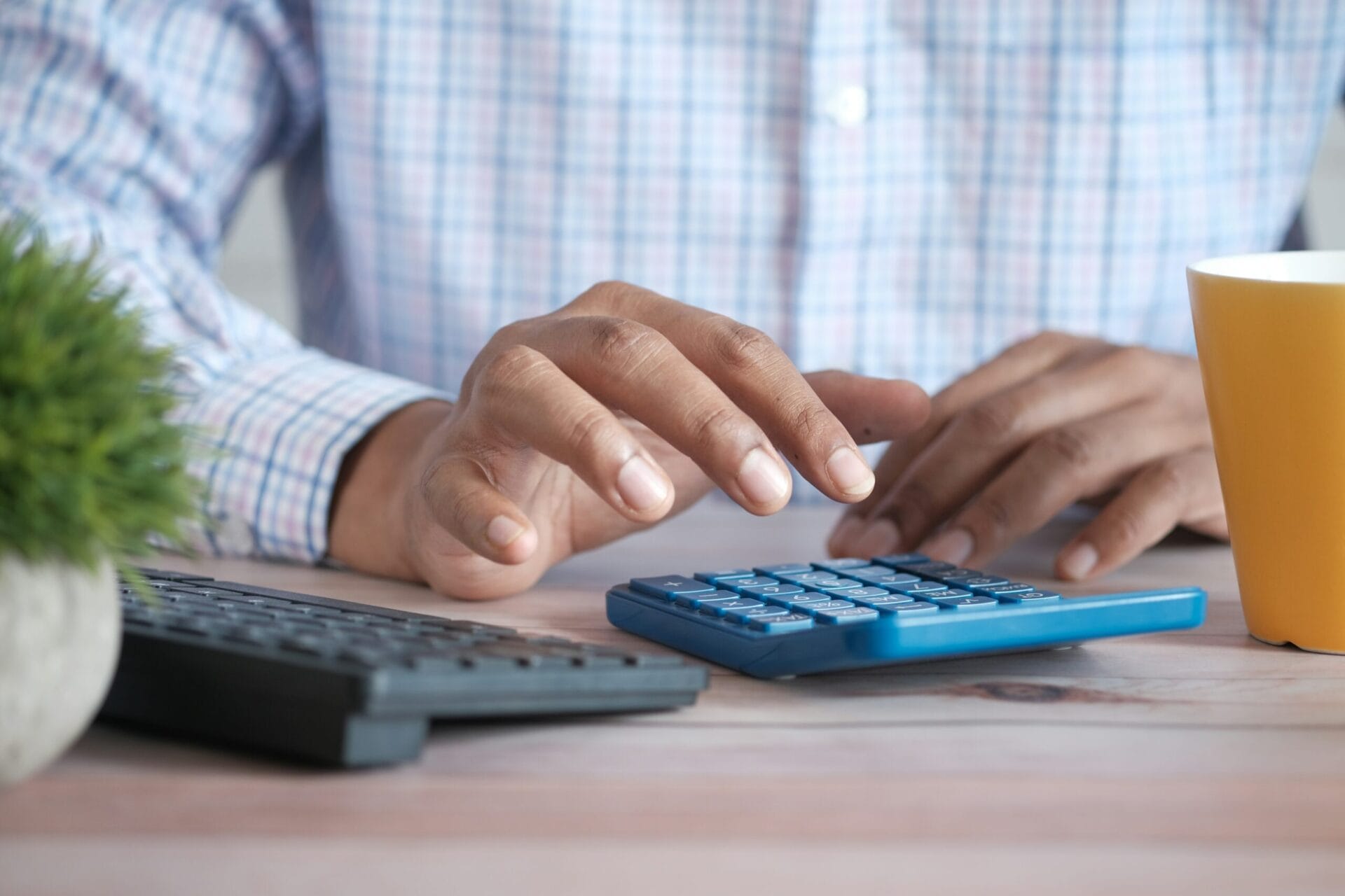 Person using a blue calculator to check holiday pay computation at a desk with a keyboard, plant, and mug.