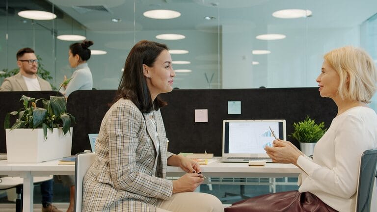 Two female coworkers in an office discussing work together at a desk, representing the equal treatment and workplace collaboration protected under the Magna Carta of Women
